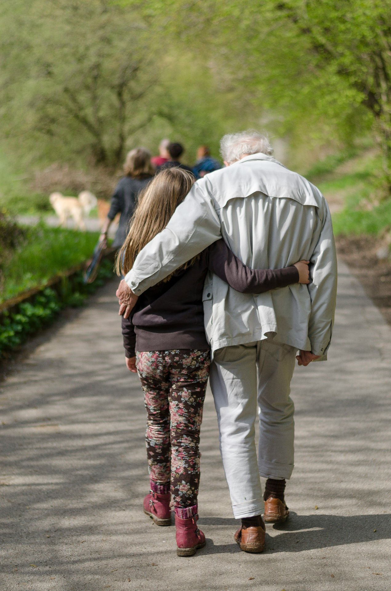 A grandparent and granddaughter walking arm in arm down a tree-lined path